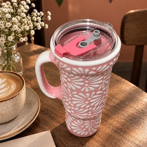 May include: A pink and white ceramic tumbler with a raised floral pattern and a handle. The tumbler features a clear plastic lid with a pink sliding closure mechanism. The word TALAVERARTE is visible on the surface of the tumbler's packaging.