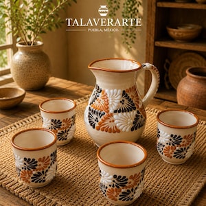Op de afbeelding: Een keramische kan en drie bijpassende bekers met een crèmekleurige basis en handgeschilderde bloemmotieven in bruine, oranje en witte tinten. De set bevat de tekst TALAVERARTE PUEBLA, MEXICO met een klein icoon van een vaas erboven.
