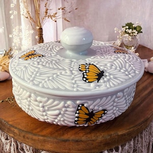 May include: A light gray ceramic dish with a lid, adorned with embossed floral patterns and several orange and black butterfly decals. The lid features a round knob. The dish sits on a wooden surface.