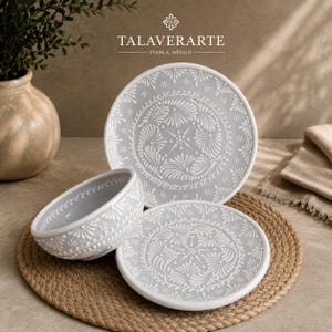 Puede incluir: Un juego de vajilla de cerámica que incluye un plato grande, un plato pequeño y un cuenco, todos en color gris claro con patrones florales y geométricos blancos en relieve. El texto TALAVERARTE PUEBLA, MEXICO aparece en la parte superior.