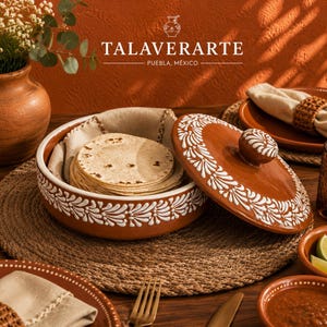 May include: A round terracotta tortilla warmer with a lid, featuring white hand-painted floral patterns. The container holds a stack of round flatbreads inside a cloth napkin. The text TALAVERARTE PUEBLA, MÉXICO is visible above the container.