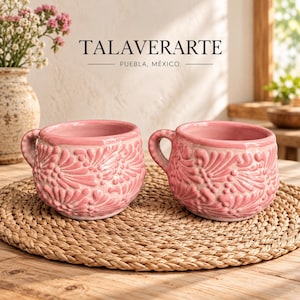 Könnte beinhalten: Zwei passende rosafarbene Keramikbecher mit einem erhabenen, geprägten Blumenmuster auf der Außenseite. Jeder Becher hat einen abgerundeten Körper und einen seitlichen Henkel. Der Text TALAVERARTE PUEBLA, MÉXICO steht schwarz über den Bechern.