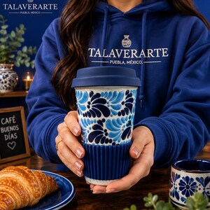 Puede incluir: Una taza de viaje de cerámica con base blanca y un patrón floral azul pintado a mano, rematada con una tapa de silicona azul oscuro y una funda de agarre de silicona acanalada a juego en la parte inferior del recipiente cilíndrico.