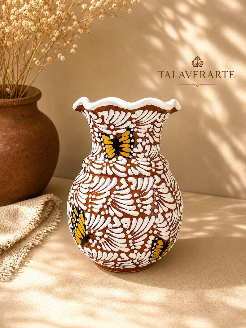 May include: A ceramic vase with a brown base colour, featuring a white scalloped rim and intricate white painted patterns covering the surface. Three yellow and black butterfly motifs are painted on the exterior. The text TALAVERARTE is visible.