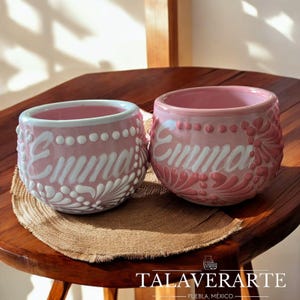 Puede incluir: Dos macetas de cerámica rosa con el nombre "Emma" en escritura blanca. Las macetas tienen un diseño floral decorativo y están sobre una estera de yute. Las macetas están sobre una mesa de madera.