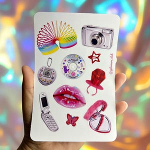 Puede incluir: Una hoja de pegatinas con varios gráficos temáticos de Y2K. Incluye un muelle en espiral arcoíris, una cámara digital, una estrella, un CD, un chupete rojo, un Tamagotchi, un teléfono plegable, labios rosas, una mariposa y un espejo compacto. El texto "@aphemera.club" es visible.