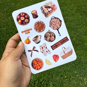 Könnte beinhalten: Ein Bogen mit herbstlichen Aufklebern mit Äpfeln, Kürbiskuchen, Zimtschnecken und einem Korb mit Kürbissen. Der Text "THE EPHEMERA CLUB" und "fresh cider" sind ebenfalls zu sehen. Die Aufkleber werden in einer Hand vor grünem Gras gehalten.