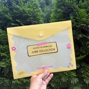 Könnte beinhalten: Eine durchscheinende gelbe Dokumentenmappe aus Kunststoff mit Druckknopfverschluss. Auf der Vorderseite befindet sich ein Etikett mit dem Text SAVING FOR LATER EVER JUNK COLLECTION, verziert mit kleinen Aufklebern von Blume, Lippen und Sternen sowie XOXO.