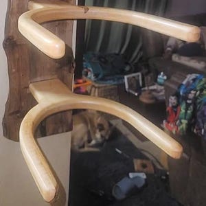 May include: A wooden wall-mounted coat rack with a natural edge, featuring two curved, light-colored wooden arms. The rack is mounted on a dark brown, textured wooden base with the name "Sanchez" engraved on it. The rack is designed to hold coats or other items.