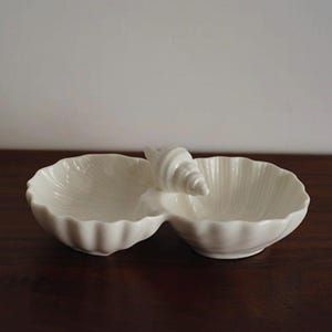 May include: A white ceramic seashell-shaped serving dish with two compartments. The dish has a textured, ridged design and a sculpted seashell handle. The dish is on a dark wooden surface.