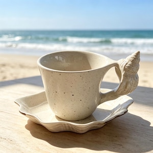 Könnte beinhalten: Ein gesprenkeltes, cremeweißes Tassen- und Untertassen-Set aus Keramik. Die Tasse hat einen Henkel in Form einer strukturierten Muschel. Die Untertasse besitzt einen gewellten, geschwungenen Rand, der an eine Muschel erinnert. Die Keramik ist glatt.
