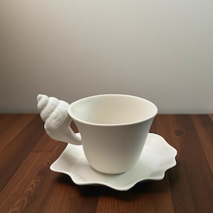Peut inclure: Un service à thé en céramique blanche. La tasse a une anse en forme de coquillage. La soucoupe a un bord festonné. L'ensemble est posé sur une surface en bois foncé.