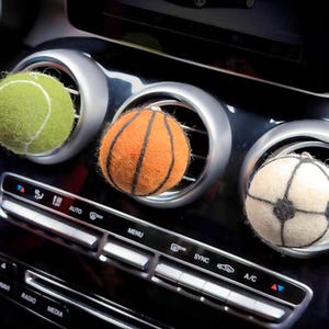 May include: Three sports ball-shaped car air fresheners. A green tennis ball, an orange basketball, and a white soccer ball with black accents are placed in car vents. The car's dashboard is black with silver air vent surrounds.