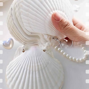 May include: A stack of white seashell-shaped notepads held by a hand, with a pearl beaded handle. The notepads are open, revealing the pages. Two iridescent heart-shaped decorations are in the background.