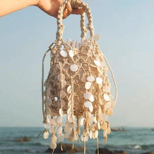 May include: A beige macrame bucket bag with white, circular embellishments. The bag has a woven handle and a drawstring closure. The bag is held up against a backdrop of the sea and sky.