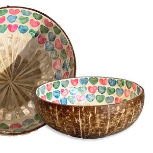 Puede incluir: Dos cuencos decorativos de coco. El cuenco más grande tiene un diseño de mosaico con formas de corazón coloridas. El cuenco más pequeño tiene un exterior marrón y un interior con estampado de corazón en tonos rosa, azul y verde.
