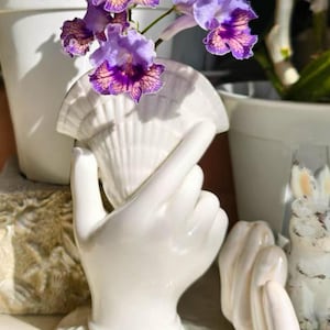 Könnte beinhalten: Eine weiße Keramikvase in Form einer Hand, die eine Muschel hält, mit violett und orange gestreiften Blumen. Der Sockel der Vase hat eine muschelartige Textur. Die Hand ist so positioniert, dass die Finger um das Muschelgefäß gewölbt sind.