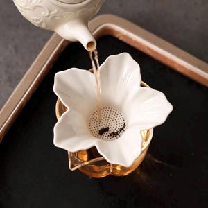 Puede incluir: Un colador de té de cerámica blanca con forma de flor de cinco pétalos y base perforada, colocado sobre una taza de cristal transparente. Una tetera de cerámica blanca con detalles en relieve vierte té a través del colador con hojas de té.