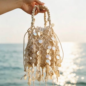May include: A beige macrame drawstring bag featuring a knotted net design adorned with numerous round, iridescent white shell-like discs that hang as fringe. The bag includes a braided handle and a solid beige fabric interior lining.