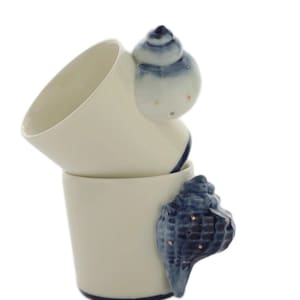 Puede incluir: Dos tazas de cerámica apiladas con un acabado liso en color blanco roto y un borde azul oscuro en la base. Cada taza presenta un asa tridimensional en forma de concha marina en tonos azules y blancos, con pequeños puntos dorados.
