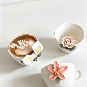 以下が含まれることがあります： 装飾的な花のアクセントが付いた3つの白いセラミックカップ。1つはスワンのラテアートと白いカラーリリーが施されたコーヒー入り。もう1つは彫刻されたピンクのバラ、3つ目は中央が黄色のピンクの花がデザインされています。