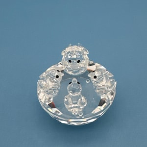 May include: A clear faceted crystal figurine featuring four small ducklings arranged in a circular formation on a round, reflective crystal base. Each duckling has tiny black eyes and a smooth, polished finish that catches the light.