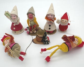 May include: A collection of seven small gnome figurines featuring felt and fabric clothing in red, yellow, and beige. The figures include standing gnomes with pointed hats and two figures lying down, one with a small sled and one with a red instrument.