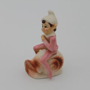 May include: A ceramic figurine of an elf in a pink outfit and white hat, seated on a snail shell. The elf has a rosy complexion and is posed with hands clasped. The snail shell is cream and brown.
