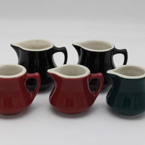 May include: Five small ceramic cream pitchers in a variety of colors. Two are black, two are red, and one is dark green. Each pitcher has a white interior and a handle.