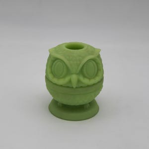 May include: A light green owl-shaped decorative item. The owl has large, oval eyes, a small beak, and textured feathers. It features a circular opening at the top and sits on a round base. The item is made of a solid, opaque material.