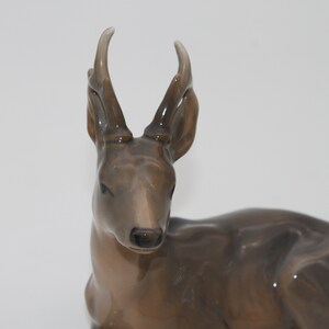 May include: A ceramic figurine of a brown deer with large antlers. The deer is lying down with its head turned to the side.