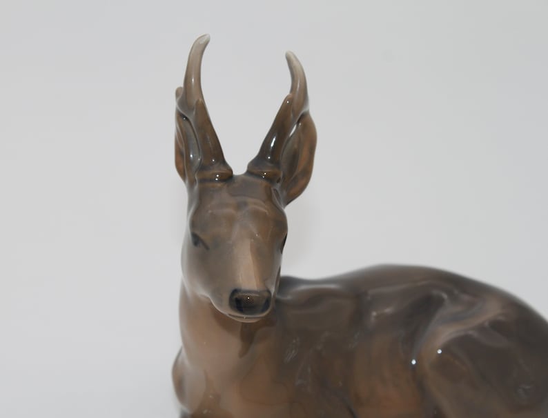 May include: A ceramic figurine of a brown deer with large antlers. The deer is lying down with its head turned to the side.