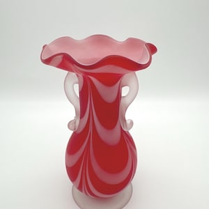 May include: A glass vase featuring a vibrant red and white swirl pattern. The vase has a wide, ruffled rim, two curved handles on the sides, and a solid, frosted base. The body of the vase displays a smooth, glossy finish with vertical, flowing lines.
