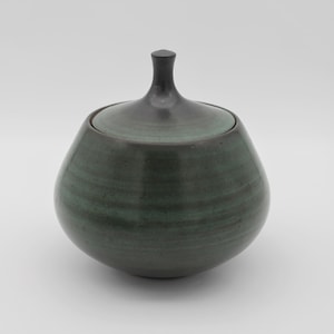 May include: A handcrafted ceramic jar with a lid. The jar is a deep teal color with darker accents and a smooth, rounded shape. The lid is a darker shade of teal with a small, rounded handle. The jar is likely used for storage or decoration.