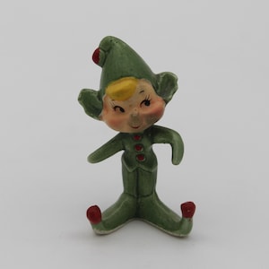 May include: A vintage ceramic elf figurine. The elf is wearing a green outfit with red accents on the hat, buttons, and shoes. The elf has a smiling face with rosy cheeks, blonde hair, and large ears. The figurine is sitting down.