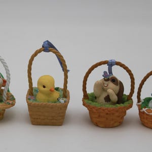 May include: Four miniature Easter baskets, each with a different animal figurine. The figurines include a dog, a duckling, a puppy with a butterfly, and a white mouse with a berry. The baskets are woven with brown handles.