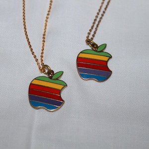 May include: Two necklaces featuring apple-shaped pendants with horizontal stripes in green, yellow, orange, red, purple, and blue enamel. Each pendant hangs from a thin gold-toned metal chain with a small green leaf detail at the top.