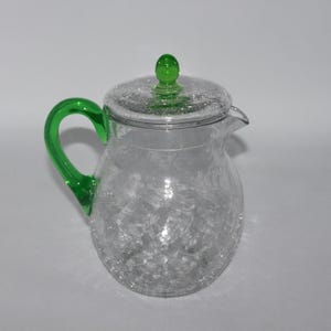 May include: A clear glass pitcher with a crackled texture, a green handle, and a matching green knob on the lid. The pitcher has a spout for easy pouring and is designed for serving beverages.