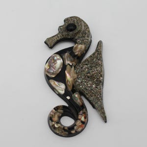 May include: A seahorse-shaped decorative item with a black base, adorned with iridescent shell pieces and textured, speckled accents. The seahorse has a curved tail and a detailed head, creating a unique coastal or nautical theme.
