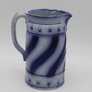 May include: A blue and white ceramic pitcher with a spiral design. The pitcher has a handle and a spout. The top and bottom of the pitcher have decorative trim. The pitcher is approximately 10 inches tall.