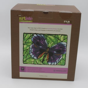May include: A box for a needlepoint kit featuring a purple butterfly with green foliage. The box is brown with the text "artiste" and "$79.99" on the front. The text "The easy way to DIY: Create a stunning design in a few simple steps, and display it wherever you please!" is also on the front of the box.