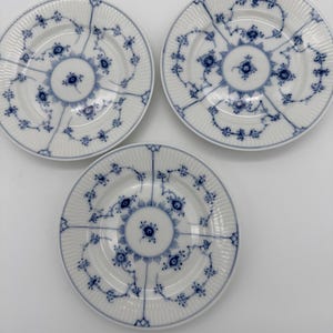 May include: Three white porcelain plates featuring a delicate blue floral pattern. Each plate has a fluted rim and a central circular motif surrounded by intricate, symmetrical blue vine and flower designs that extend toward the edges.