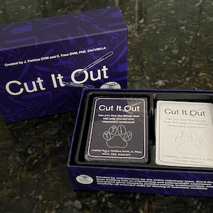 Puede incluir: Un juego de mesa llamado "Cut It Out" con una caja y cartas moradas. El título del juego está en blanco en la caja y las cartas. Las cartas presentan una huella de pata y texto sobre medicina veterinaria.