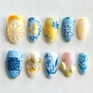 Puede incluir: Una colección de diez uñas artificiales con varios diseños. Las uñas presentan patrones florales, de limón y geométricos en tonos amarillos, azules y blancos. Algunas uñas tienen adornos 3D, incluyendo una flor y una abeja.