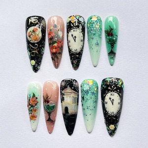 May include: A collection of ten long, almond-shaped acrylic nails with various designs. The nails feature floral patterns, clock faces, and glitter accents in colors like black, pink, green, and white. Some nails have the number "26" on them.