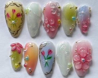 Könnte beinhalten: Ein Set aus zehn mandelförmigen Kunstnägeln mit verschiedenen floralen Designs. Die Nägel weisen eine Reihe von Farben auf, darunter Rosa, Gelb und Blau, mit goldenen Akzenten und Perlenverzierungen. Einige Nägel haben 3D-Blumendesigns.