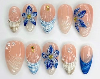 May include: A set of ten artificial nails with a variety of designs. The nails feature a light pink base with blue floral accents, gold embellishments, and plaid patterns. Some nails have a white and blue swirl design.