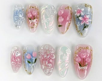 Op de afbeelding: Een set van tien kunstnagels met diverse ontwerpen. De nagels hebben een reeks kleuren, waaronder roze, blauw en wit, met gouden accenten. Ontwerpen omvatten bloempatronen, strikken en abstracte vormen. De nagels zijn ovaal van vorm.
