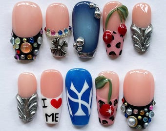Puede incluir: Un conjunto de diez uñas artificiales con varios diseños. Algunas uñas son nude con adornos, incluyendo pedrería y detalles plateados. Otras uñas presentan un degradado azul, un diseño de cereza y el texto "I ❤️ ME".