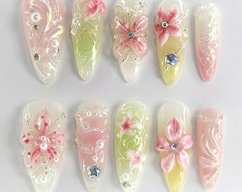 May include: Ten long, oval-shaped artificial nails with a white base and pink floral designs. Some nails have silver chains and pearl accents.